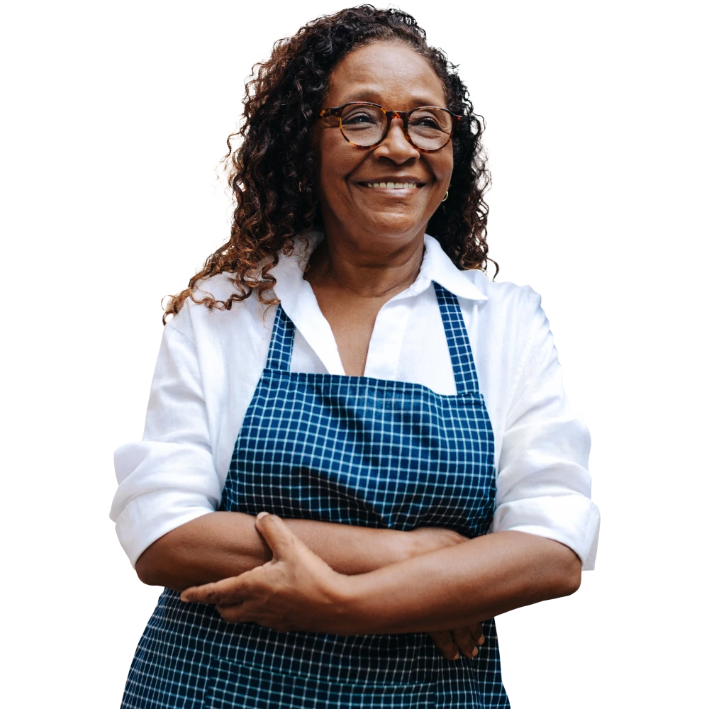 Smiling older woman wearing glasses and a blue checkered apron with arms crossed
