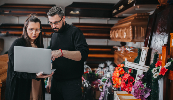 Two people discuss options over a laptop in a funeral home with caskets and crosses.